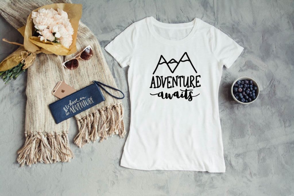 White 'Adventure Awaits' t-shirt styled with sunglasses, flowers, and a wristlet