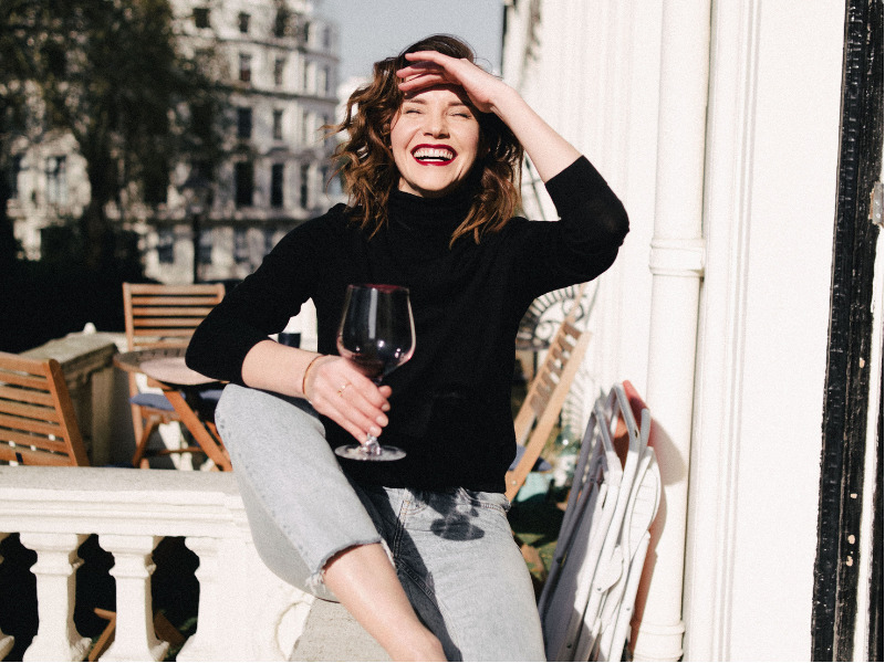 Woman with a glass of red wine laughing in the sun on a balcony