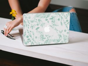 Laptop decorated with a green floral decal on a table beside a pair of glasses and a small vase of flowers