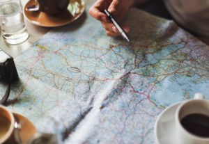 Hand holding a pen over a paper map next to a cup of coffee and a camera