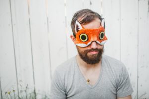 Bearded man wearing an orange felt fox sleeping mask against a wooden fence