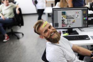 Bearded man laughing at his desk with a yellow sticky note stuck to his forehead