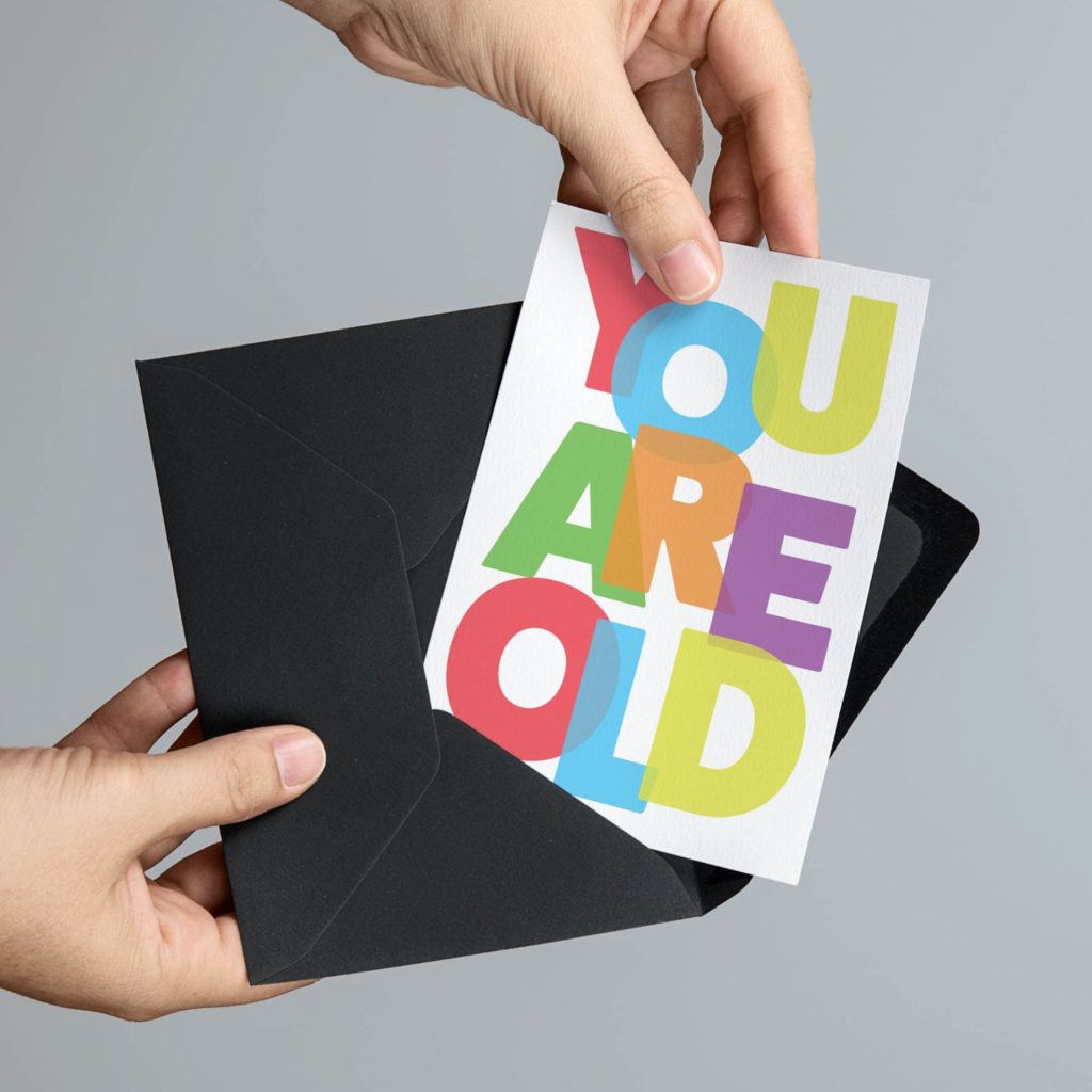 Two hands sliding a colorful 'You Are Old' greeting card into a black envelope